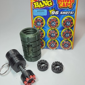 May include: A selection of toy items, including a green grenade-shaped object, a black toy gun with a spring, and two black ring caps. A package of "RING CAPS" with 96 shots is also present. The packaging has the text "AGE 12+".