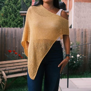 Puede incluir: Un poncho de punto amarillo mostaza con un dise&ntilde;o asim&eacute;trico. El poncho est&aacute; drapeado sobre los hombros, revelando una parte superior blanca debajo. La modelo lleva vaqueros azul oscuro. El fondo incluye una valla de madera y vegetaci&oacute;n.