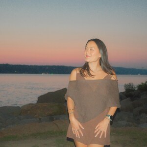 Puede incluir: Una persona con un vestido marr&oacute;n con los hombros descubiertos est&aacute; de pie cerca de una masa de agua al anochecer. El cielo pasa del rosa al azul. El vestido tiene un chal a juego. La persona lleva joyas y un reloj.