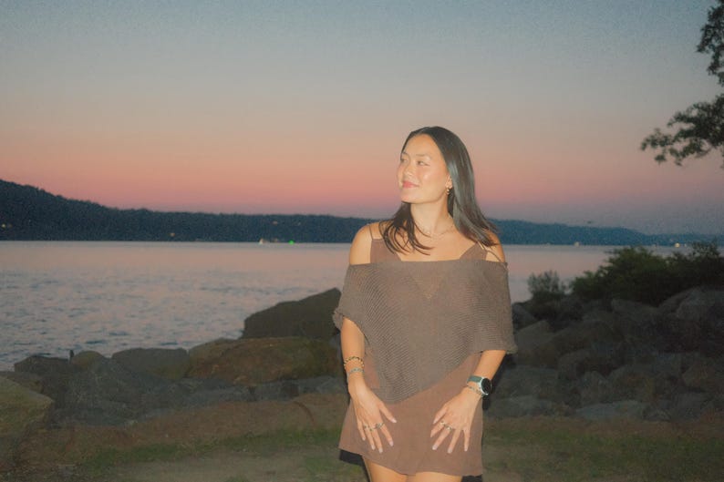 Puede incluir: Una persona con un vestido marr&oacute;n con los hombros descubiertos est&aacute; de pie cerca de una masa de agua al anochecer. El cielo pasa del rosa al azul. El vestido tiene un chal a juego. La persona lleva joyas y un reloj.