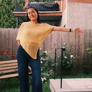 Puede incluir: Una mujer con un top estilo poncho amarillo mostaza y vaqueros azul oscuro posa al aire libre. El top tiene un dise&ntilde;o suelto. El fondo incluye una valla de madera, un banco y vegetaci&oacute;n.