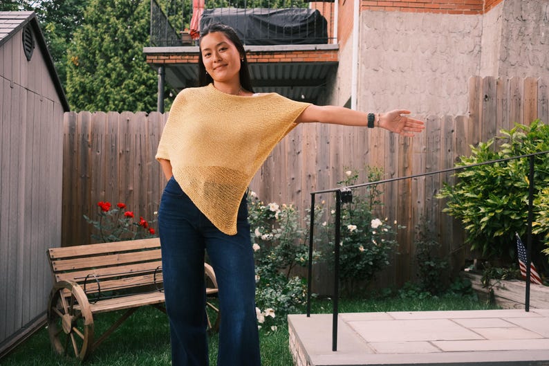Puede incluir: Una mujer con un top estilo poncho amarillo mostaza y vaqueros azul oscuro posa al aire libre. El top tiene un dise&ntilde;o suelto. El fondo incluye una valla de madera, un banco y vegetaci&oacute;n.