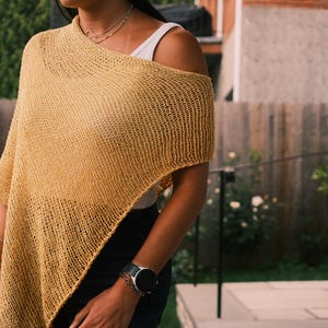 Puede incluir: Un poncho tejido de color amarillo mostaza con un dise&ntilde;o asim&eacute;trico. El poncho est&aacute; hecho de una tela texturizada de tejido abierto. El modelo lleva una camiseta sin mangas blanca debajo y un collar plateado.