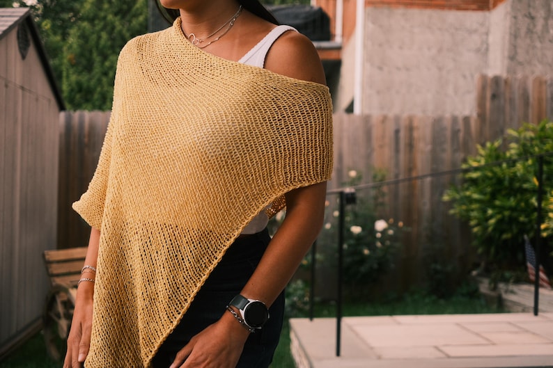 Puede incluir: Un poncho tejido de color amarillo mostaza con un dise&ntilde;o asim&eacute;trico. El poncho est&aacute; hecho de una tela texturizada de tejido abierto. El modelo lleva una camiseta sin mangas blanca debajo y un collar plateado.