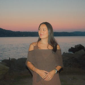 May include: A woman wearing a brown off-the-shoulder top and dress, standing near a body of water at dusk. The sky transitions from pink to blue, with a dark mountain range in the background. She is wearing a watch and bracelets.