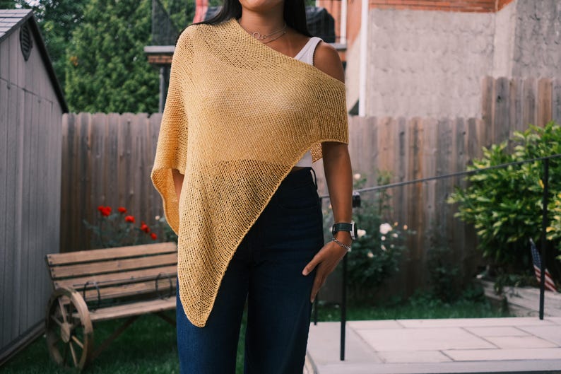 Puede incluir: Un poncho de punto amarillo mostaza con un dise&ntilde;o asim&eacute;trico. El poncho est&aacute; drapeado sobre los hombros, revelando una parte superior blanca debajo. La modelo lleva vaqueros azul oscuro. El fondo incluye una valla de madera y vegetaci&oacute;n.