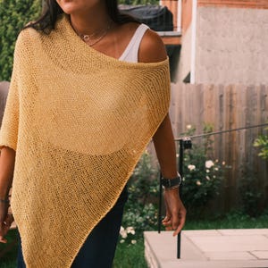 Puede incluir: Un poncho de punto amarillo mostaza con un dise&ntilde;o drapeado. La prenda de tejido abierto se lleva sobre una camiseta blanca. El poncho tiene un ajuste relajado y es adecuado para usar en capas. El fondo incluye una valla de madera y vegetaci&oacute;n.