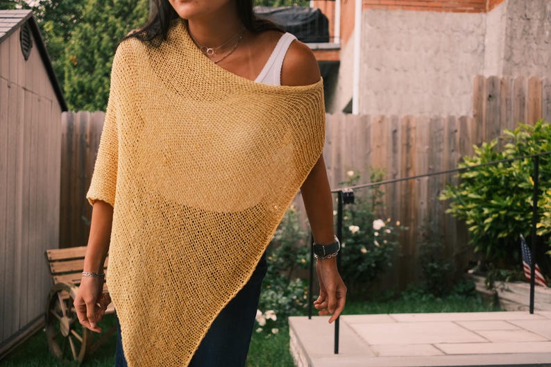 Puede incluir: Un poncho de punto amarillo mostaza con un dise&ntilde;o drapeado. La prenda de tejido abierto se lleva sobre una camiseta blanca. El poncho tiene un ajuste relajado y es adecuado para usar en capas. El fondo incluye una valla de madera y vegetaci&oacute;n.