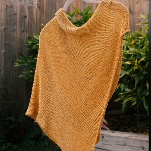 Puede incluir: Un poncho tejido de color amarillo mostaza cuelga de una percha blanca. El poncho est&aacute; hecho de una tela texturizada de tejido abierto. La prenda se exhibe al aire libre sobre un fondo de vegetaci&oacute;n y una valla de madera.