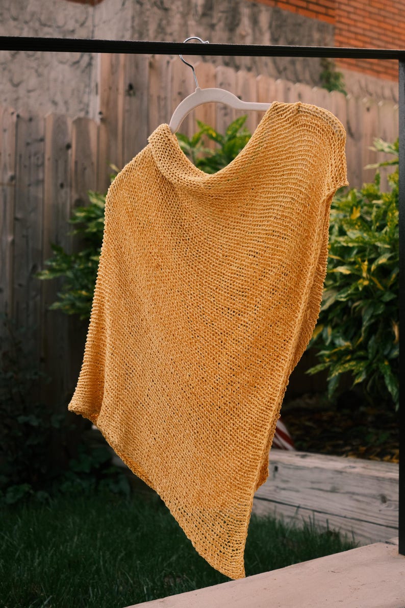 Puede incluir: Un poncho tejido de color amarillo mostaza cuelga de una percha blanca. El poncho est&aacute; hecho de una tela texturizada de tejido abierto. La prenda se exhibe al aire libre sobre un fondo de vegetaci&oacute;n y una valla de madera.