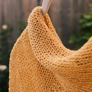 Puede incluir: Primer plano de una prenda de punto amarillo mostaza, posiblemente una bufanda o chal, colgada de una percha de madera. El tejido tiene un aspecto texturizado con puntadas visibles. El fondo est&aacute; borroso, lo que sugiere un entorno al aire libre.