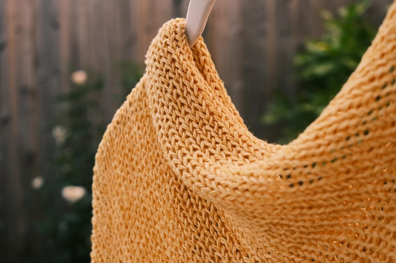 Puede incluir: Primer plano de una prenda de punto amarillo mostaza, posiblemente una bufanda o chal, colgada de una percha de madera. El tejido tiene un aspecto texturizado con puntadas visibles. El fondo est&aacute; borroso, lo que sugiere un entorno al aire libre.