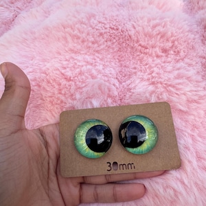 May include: Two glass doll eyes with a green and black design, each measuring 30mm. The eyes are displayed on a brown cardboard backing, held against a pink, fluffy background.