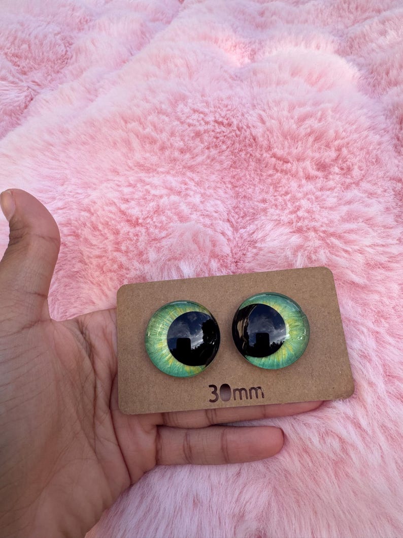 May include: Two glass doll eyes with a green and black design, each measuring 30mm. The eyes are displayed on a brown cardboard backing, held against a pink, fluffy background.