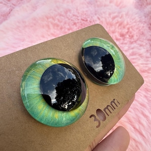 May include: Two glass eye cabochons with a green and yellow iris and a black pupil. The eyes are on a brown cardboard surface. The text "30mm" is visible on the cardboard.