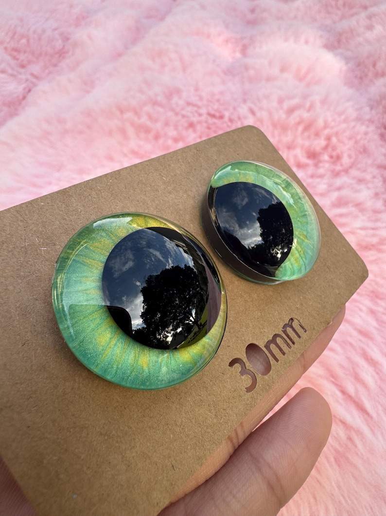May include: Two glass eye cabochons with a green and yellow iris and a black pupil. The eyes are on a brown cardboard surface. The text "30mm" is visible on the cardboard.