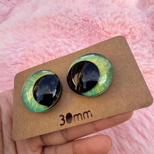 May include: Two glass doll eyes with a green iris and black pupil, set on a brown cardboard backing. The eyes are 30mm in diameter. The background is a soft, pink, fluffy material.