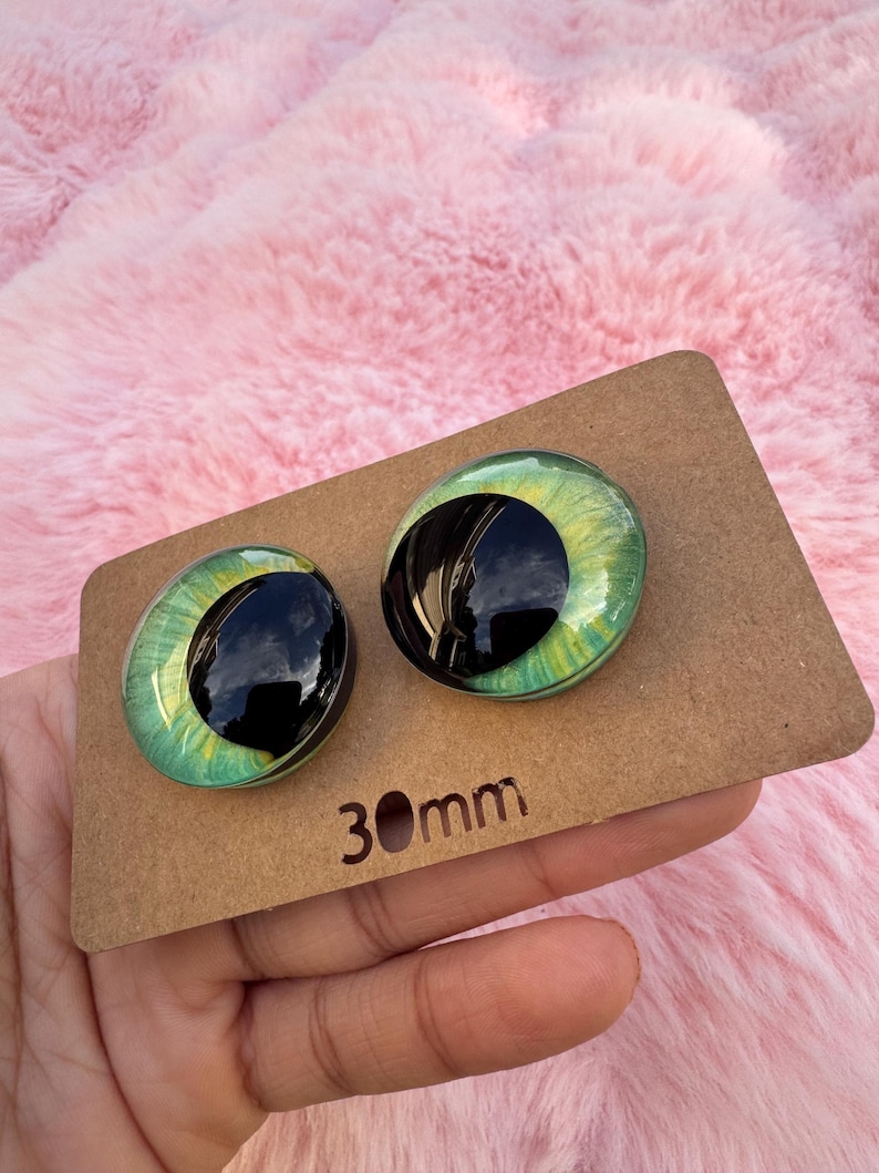 May include: Two glass doll eyes with a green iris and black pupil, set on a brown cardboard backing. The eyes are 30mm in diameter. The background is a soft, pink, fluffy material.
