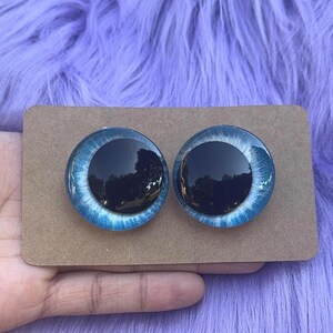 May include: Two round, blue and black craft eyes on a brown cardboard backing. The eyes have a dark center and a light blue iris with white detailing. The background is a fuzzy, purple surface.