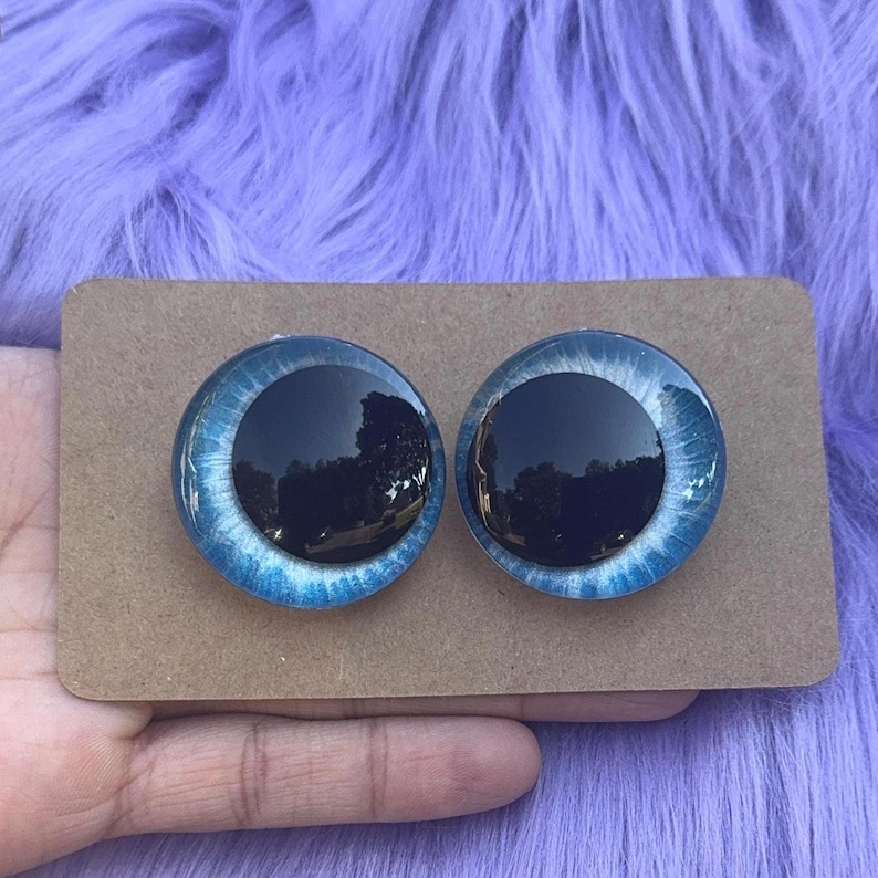 May include: Two round, blue and black craft eyes on a brown cardboard backing. The eyes have a dark center and a light blue iris with white detailing. The background is a fuzzy, purple surface.
