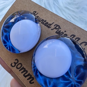 May include: Two round, blue and white plugs with a swirling blue design and a white center. The plugs are 30mm in size and are displayed on a brown card. The text "Haunted Pixie" is visible.