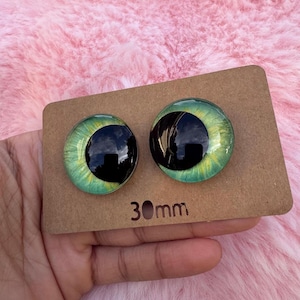 May include: Two glass eye cabochons with black pupils and green and yellow irises, displayed on a brown card. The card has the measurement "30mm" printed on it. The eyes are round and have a reflective surface.