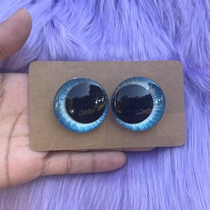 May include: Two glass doll eyes with a blue iris and black pupil, set on a brown cardboard backing. The eyes have a reflective quality, with a light blue and white pattern around the dark center. The background is a fluffy, lavender-colored material.