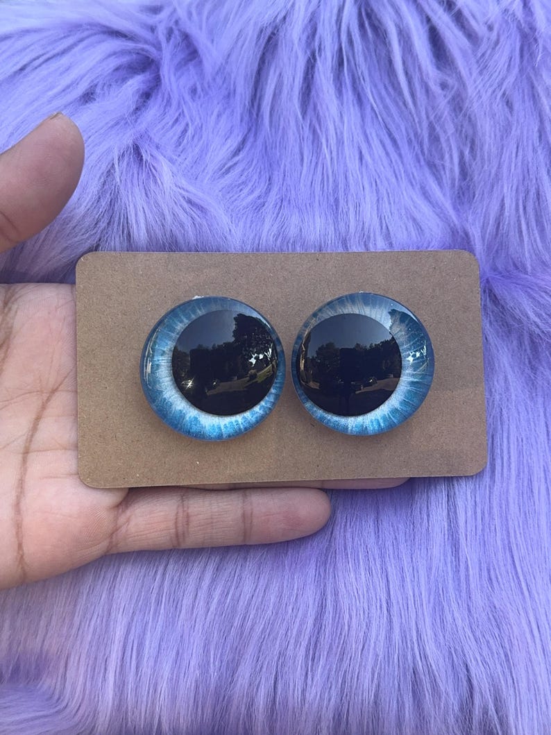 May include: Two glass doll eyes with a blue iris and black pupil, set on a brown cardboard backing. The eyes have a reflective quality, with a light blue and white pattern around the dark center. The background is a fluffy, lavender-colored material.