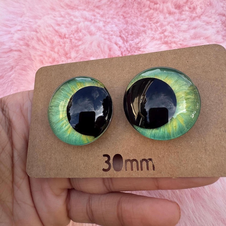 May include: Two glass doll eyes with a black pupil and green and yellow iris, each 30mm in diameter, are displayed on a brown cardboard backing. The eyes have a reflective, glossy finish, and are set against a pink, fuzzy background.