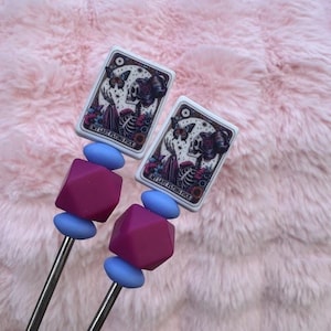 May include: Two metal skewers with decorative beads and card-shaped toppers. The toppers feature a skeleton design with butterflies and the text "MY LAST FLYING FUCK". The beads are blue and magenta, set against a pink, fluffy background.