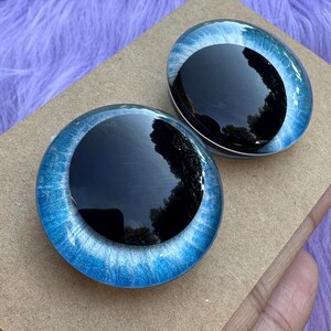 May include: Two glass doll eyes with a black iris and a blue and white outer ring. The eyes are oval-shaped and sit on a brown cardboard surface, with a purple, fuzzy background.