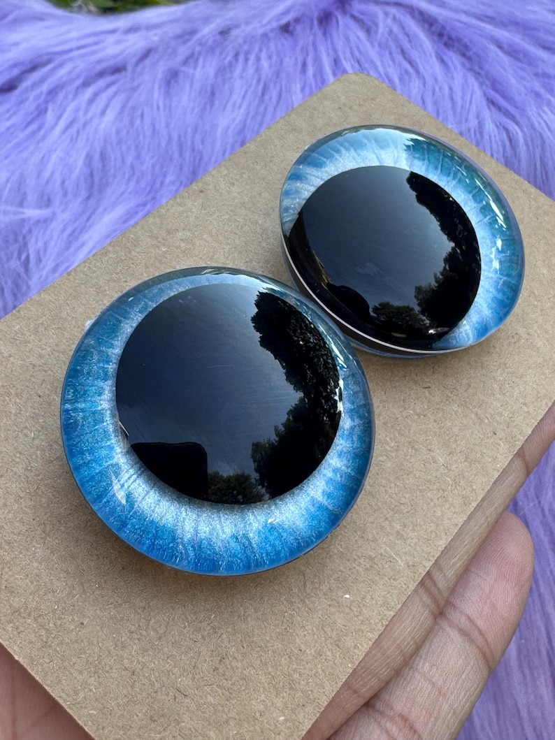May include: Two glass doll eyes with a black iris and a blue and white outer ring. The eyes are oval-shaped and sit on a brown cardboard surface, with a purple, fuzzy background.
