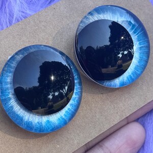 May include: Two round, blue and black doll eyes. The outer rim is a bright blue with silver glitter, and the center is a dark, reflective black. The eyes are set on a brown surface.