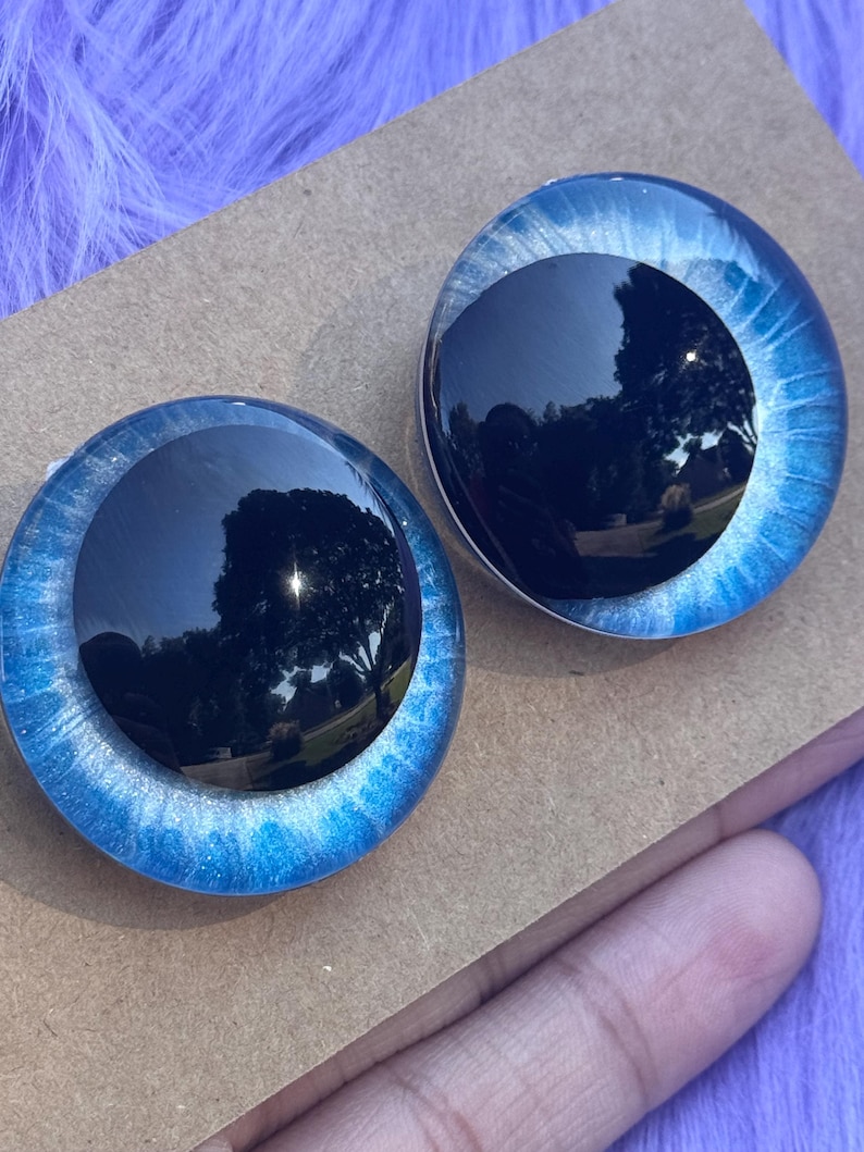 May include: Two round, blue and black doll eyes. The outer rim is a bright blue with silver glitter, and the center is a dark, reflective black. The eyes are set on a brown surface.
