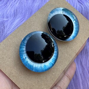 May include: Two glass doll eyes with a black pupil and a blue iris. The irises have a shimmering, textured appearance. The eyes are set on a brown cardboard surface, with a purple, fuzzy background.