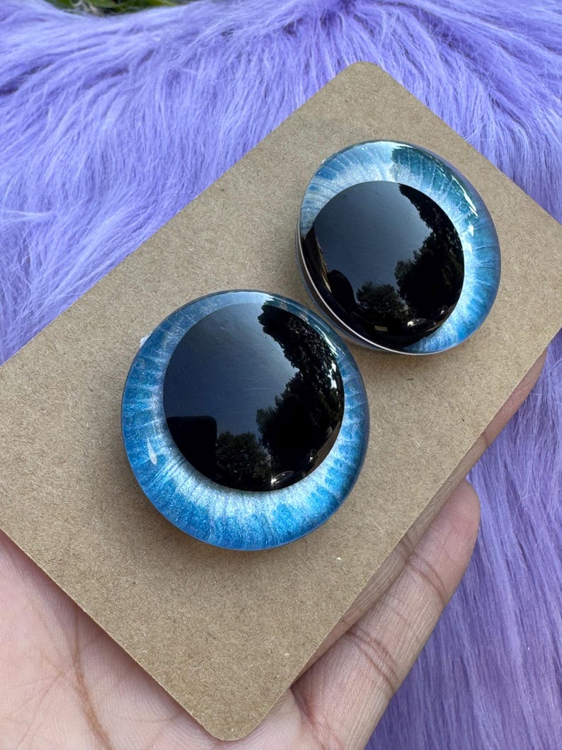 May include: Two glass doll eyes with a black pupil and a blue iris. The irises have a shimmering, textured appearance. The eyes are set on a brown cardboard surface, with a purple, fuzzy background.