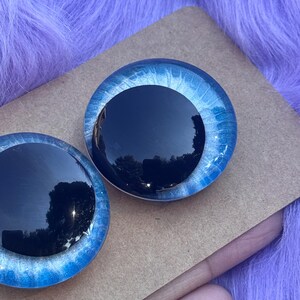 May include: Two round, glass doll eyes with a black iris and a blue and white outer ring. The eyes are set on a brown surface against a purple, fuzzy background. The eyes are reflective.