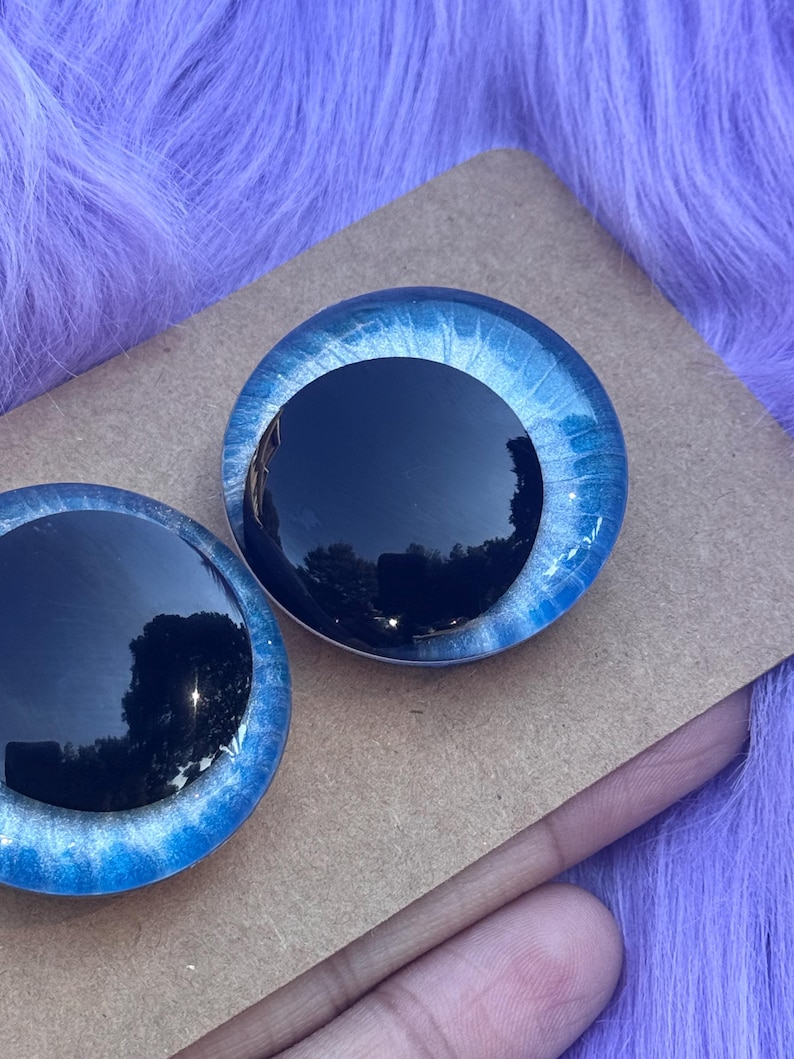 May include: Two round, glass doll eyes with a black iris and a blue and white outer ring. The eyes are set on a brown surface against a purple, fuzzy background. The eyes are reflective.