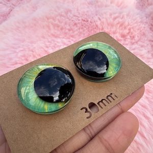 May include: Two round glass doll eyes with a black pupil and green iris, resting on a brown cardboard surface. The eyes are 30mm in diameter. The background is a soft, pink, fuzzy material.