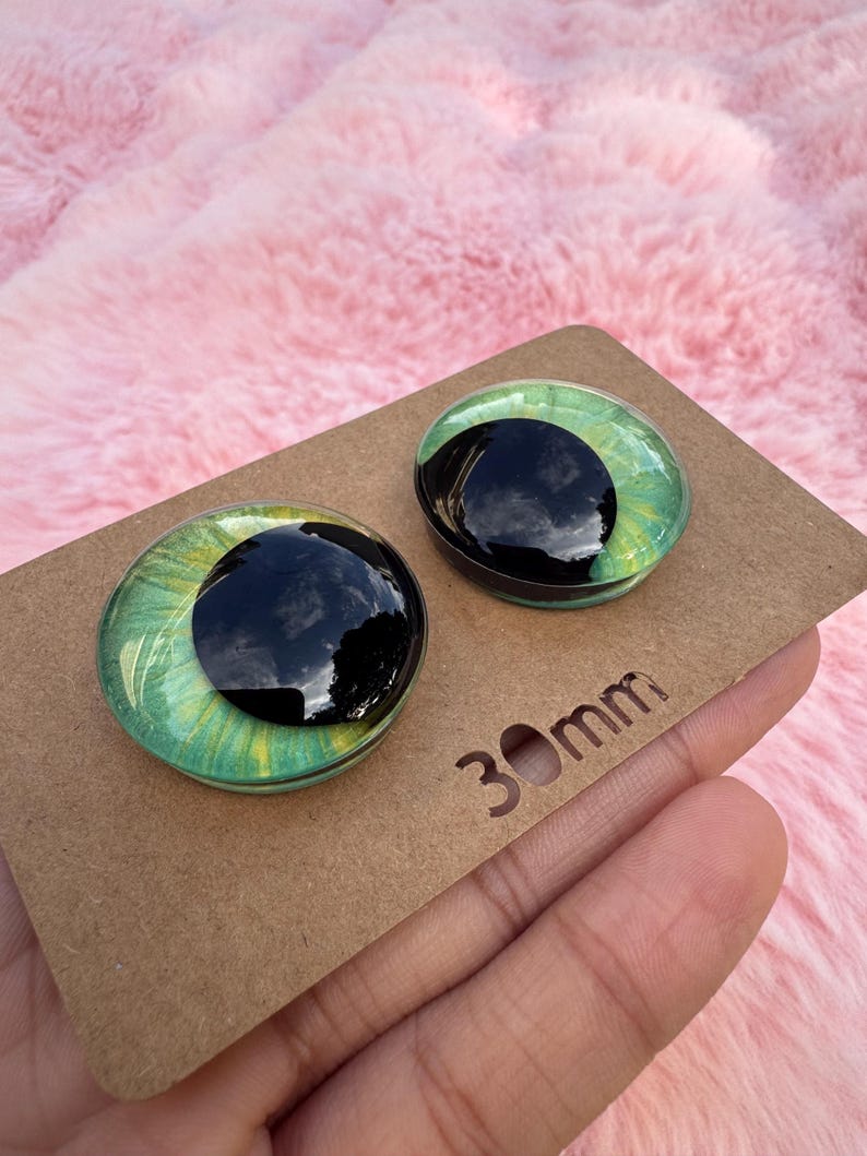 May include: Two round glass doll eyes with a black pupil and green iris, resting on a brown cardboard surface. The eyes are 30mm in diameter. The background is a soft, pink, fuzzy material.