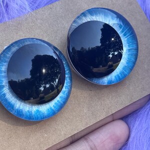 May include: Two round, blue glass doll eyes with a black pupil and a light blue and white iris. The eyes are set on a brown cardboard surface, with a purple fuzzy background.