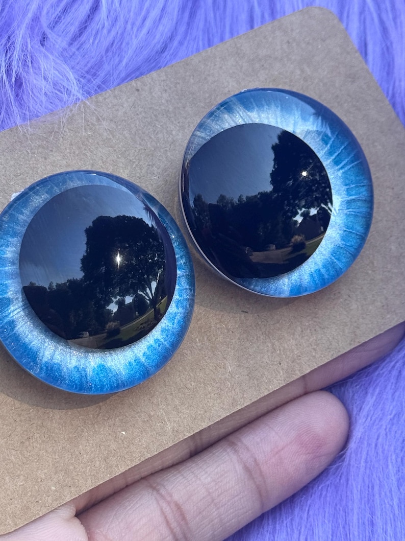 May include: Two round, blue glass doll eyes with a black pupil and a light blue and white iris. The eyes are set on a brown cardboard surface, with a purple fuzzy background.