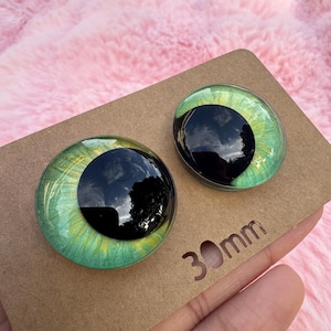 May include: Two glass doll eyes with a green iris and black pupil, set on a brown cardboard backing. The eyes are 30mm in diameter. The background is a soft pink, fluffy material.