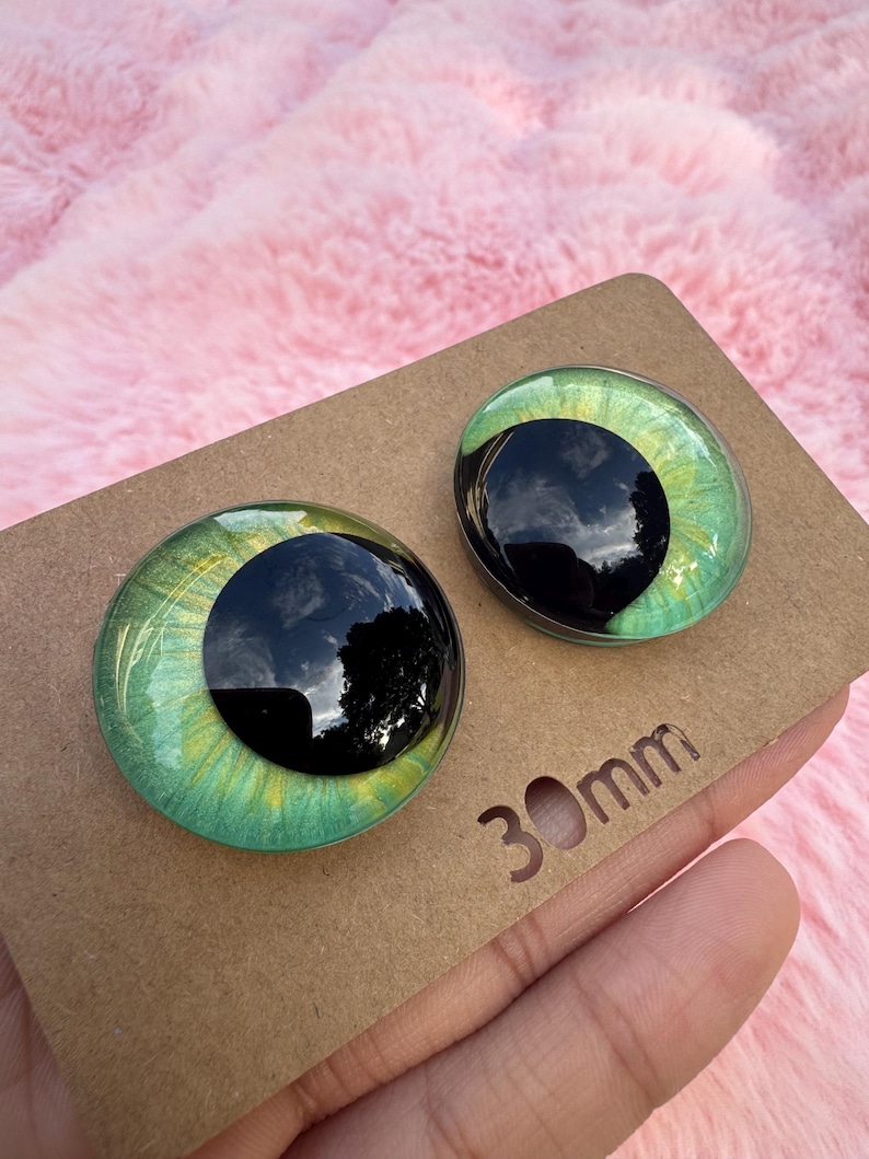 May include: Two glass doll eyes with a green iris and black pupil, set on a brown cardboard backing. The eyes are 30mm in diameter. The background is a soft pink, fluffy material.