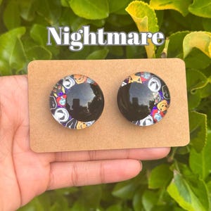 May include: Two round stud earrings with a black center and a colorful border featuring characters from "The Nightmare Before Christmas." The word "Nightmare" is above the earrings.