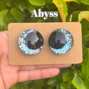 May include: A pair of round stud earrings with a black center and iridescent glitter accents. The earrings are displayed on a brown card, with the word "Abyss" in white script at the top.