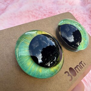 May include: Two glass eye cabochons with a black pupil and green and yellow iris. The eyes are 30mm in diameter and sit on a brown cardboard surface.