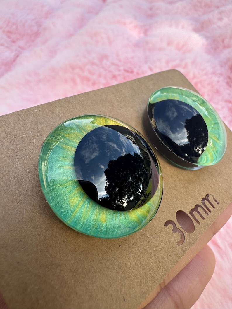 May include: Two glass eye cabochons with a black pupil and green and yellow iris. The eyes are 30mm in diameter and sit on a brown cardboard surface.