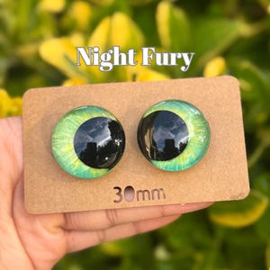 Peut inclure: Deux cabochons en verre représentant des yeux, avec une pupille noire et un iris vert, étiquetés "Night Fury". Chaque œil mesure 30 mm de diamètre. Les yeux sont présentés sur un support en carton brun.