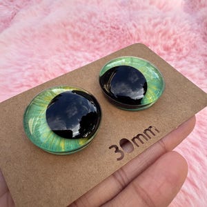 May include: Two round glass doll eyes with a black pupil and green iris, resting on a brown cardboard surface. The eyes are 30mm in diameter. The background is a soft, pink, fuzzy material.